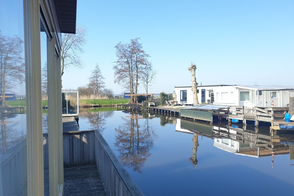 A calm water body reflecting buildings and trees, with a wooden dock and a modern house in the background.