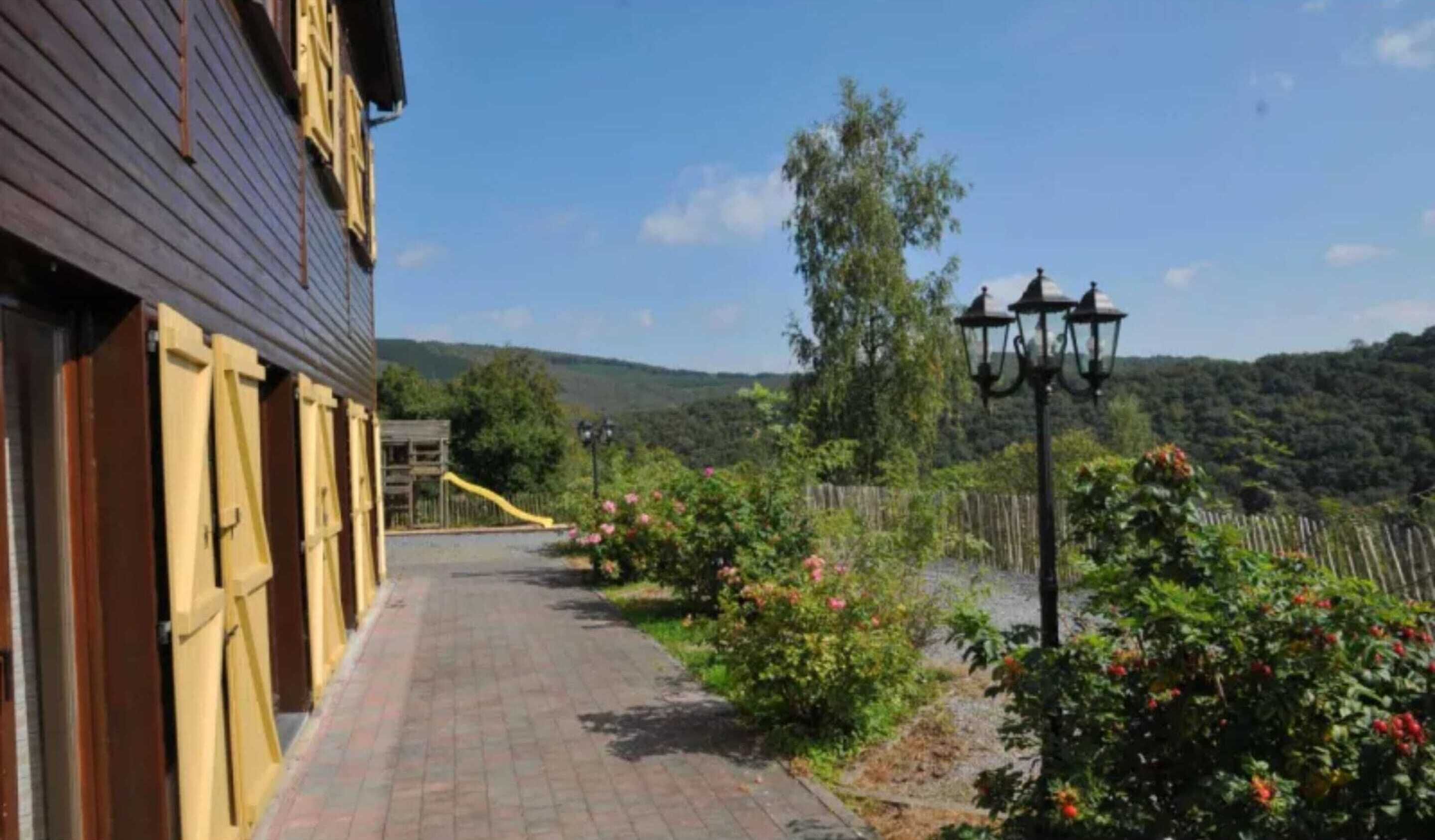 A pathway leading to a building with a wooden facade, surrounded by lush greenery and a clear sky.