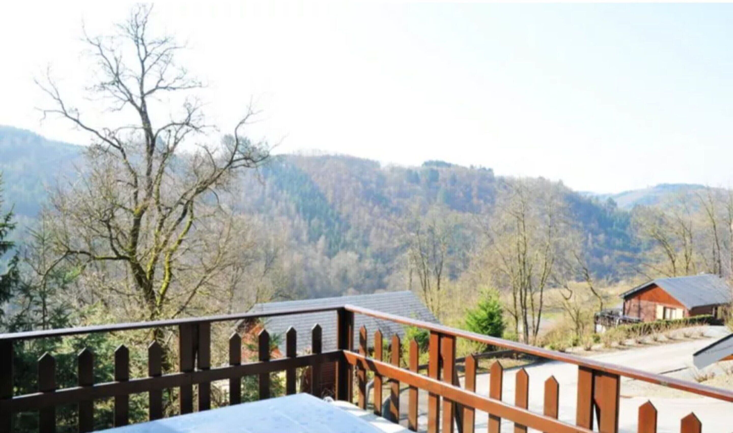 A view from a balcony with a wooden railing, overlooking a valley with trees and a mountain range in the background.
