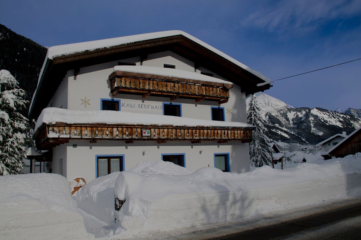 Un immeuble à plusieurs étages, dont le toit et les balcons sont recouverts de neige, entouré de neige et de montagnes.