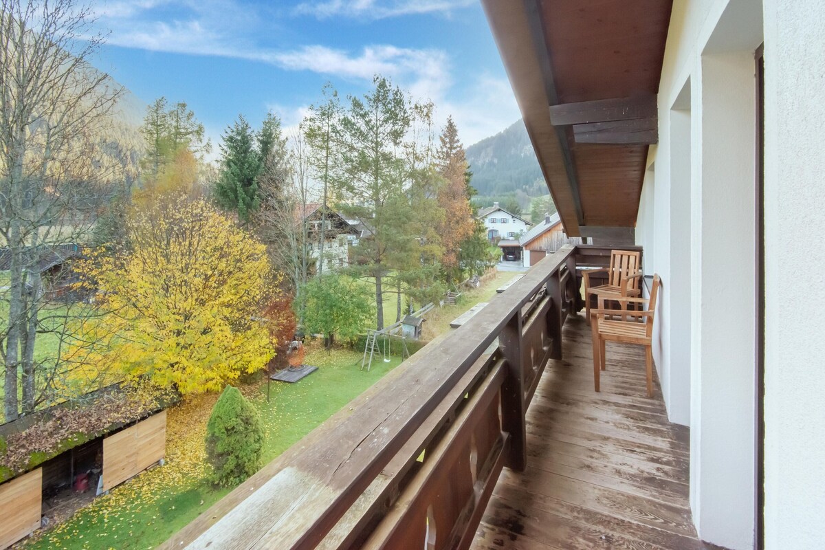 Un balcon doté d’un plancher en bois, donnant sur un jardin avec des arbres et une maison en arrière-plan.
