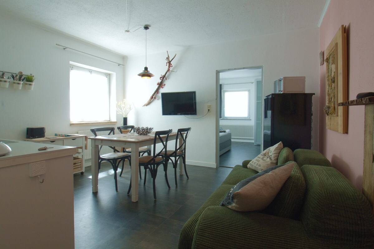 Un salon avec un canapé vert, une table à manger avec des chaises, un téléviseur fixé au mur et un coin cuisine visible à travers une porte ouverte.