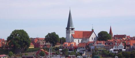 Une ville côtière qui se distingue par son imposant clocher d’église, ses bâtiments aux toits rouges et son port de plaisance où sont amarrés des bateaux.
