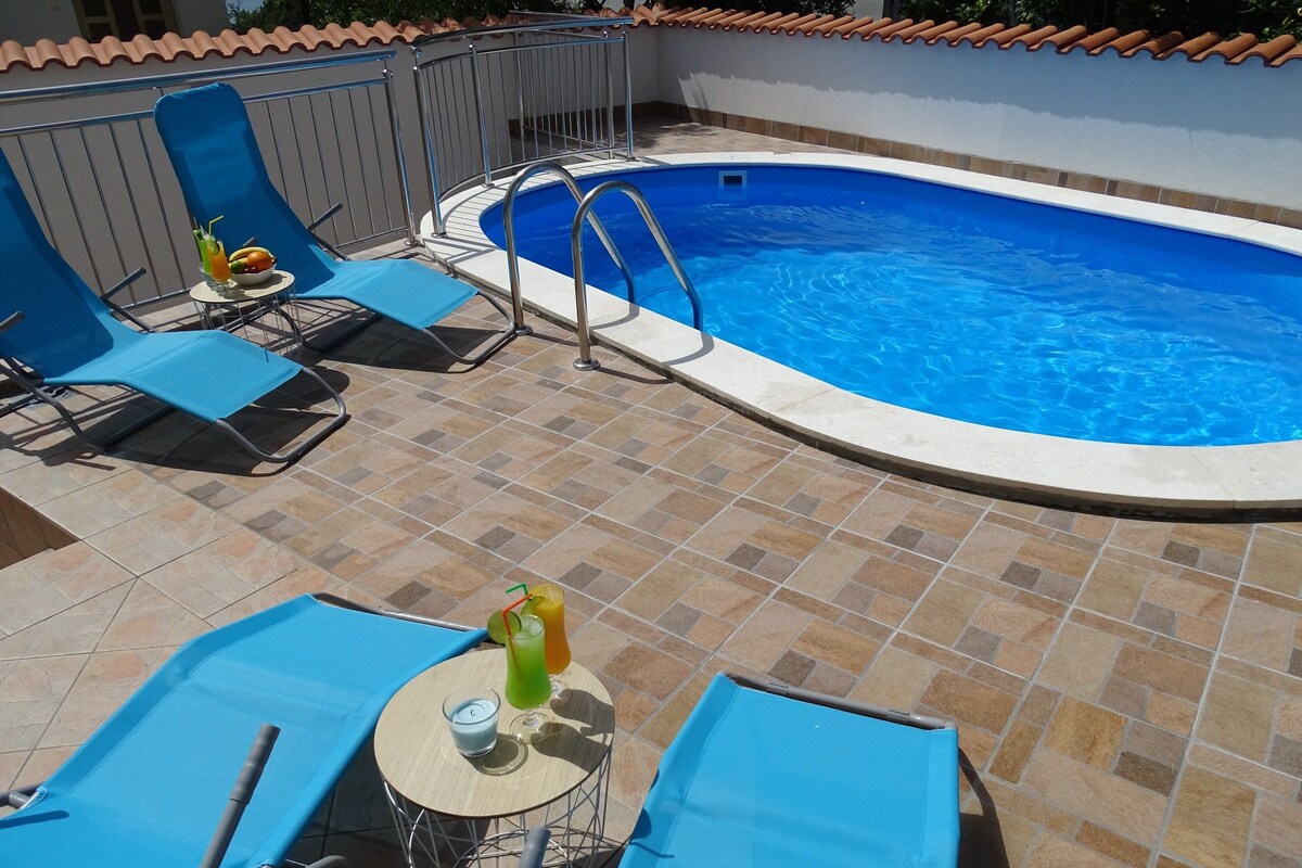 Una piscina con agua azul, rodeada de sillas reclinables azules y un solárium con baldosas.