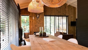 A modern dining area with a wooden table, white chairs, and a large window with blinds. - Lush Holiday Home in Rheezerveen With Hot Tub (Rheezerveen)
