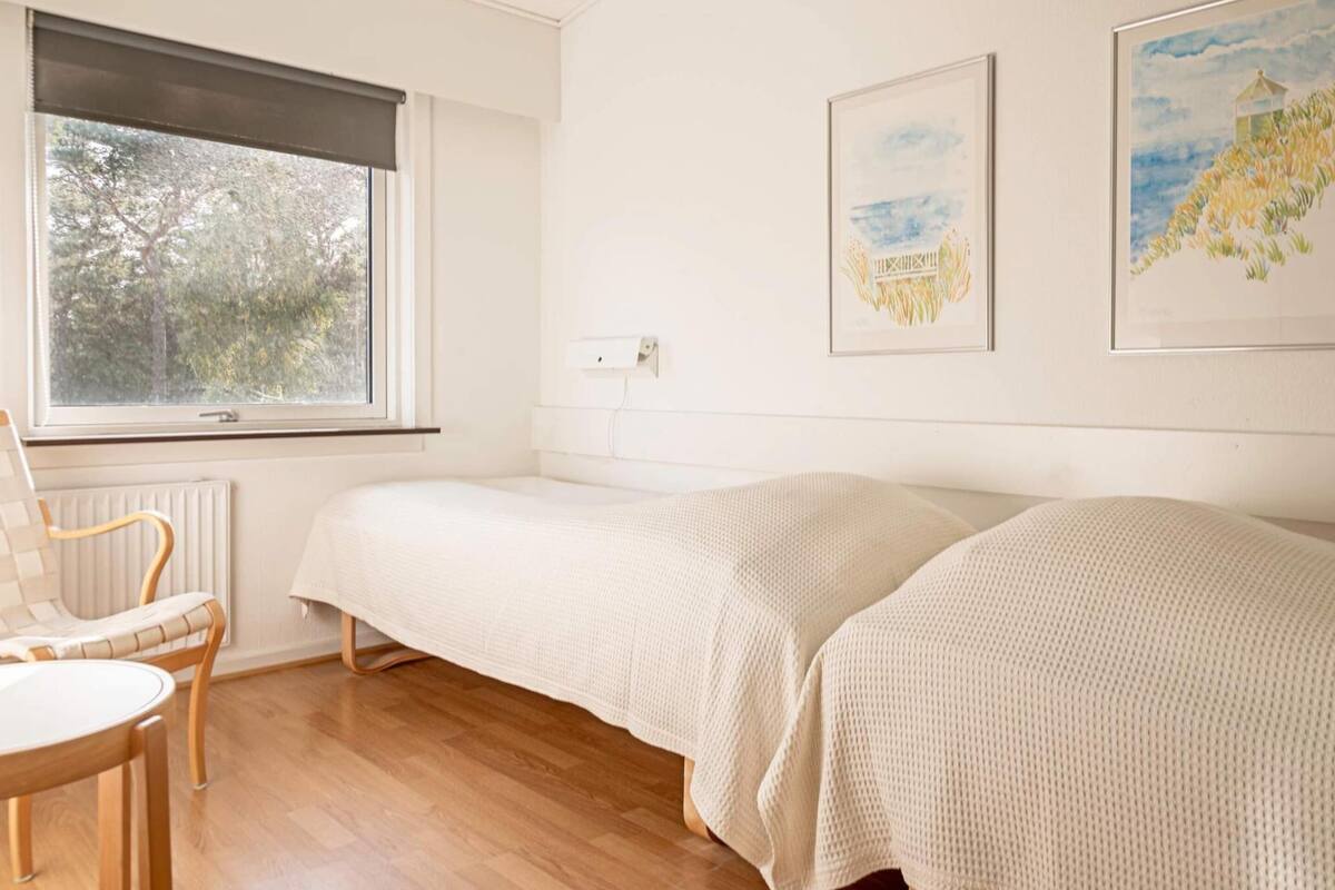 A room with two single beds, a chair, a radiator, and a window with a view of trees.