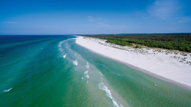 Ett flygbildsbild av en strand med kristallklart turkosfärgat vatten, vit sand och ett skogsområde i bakgrunden.