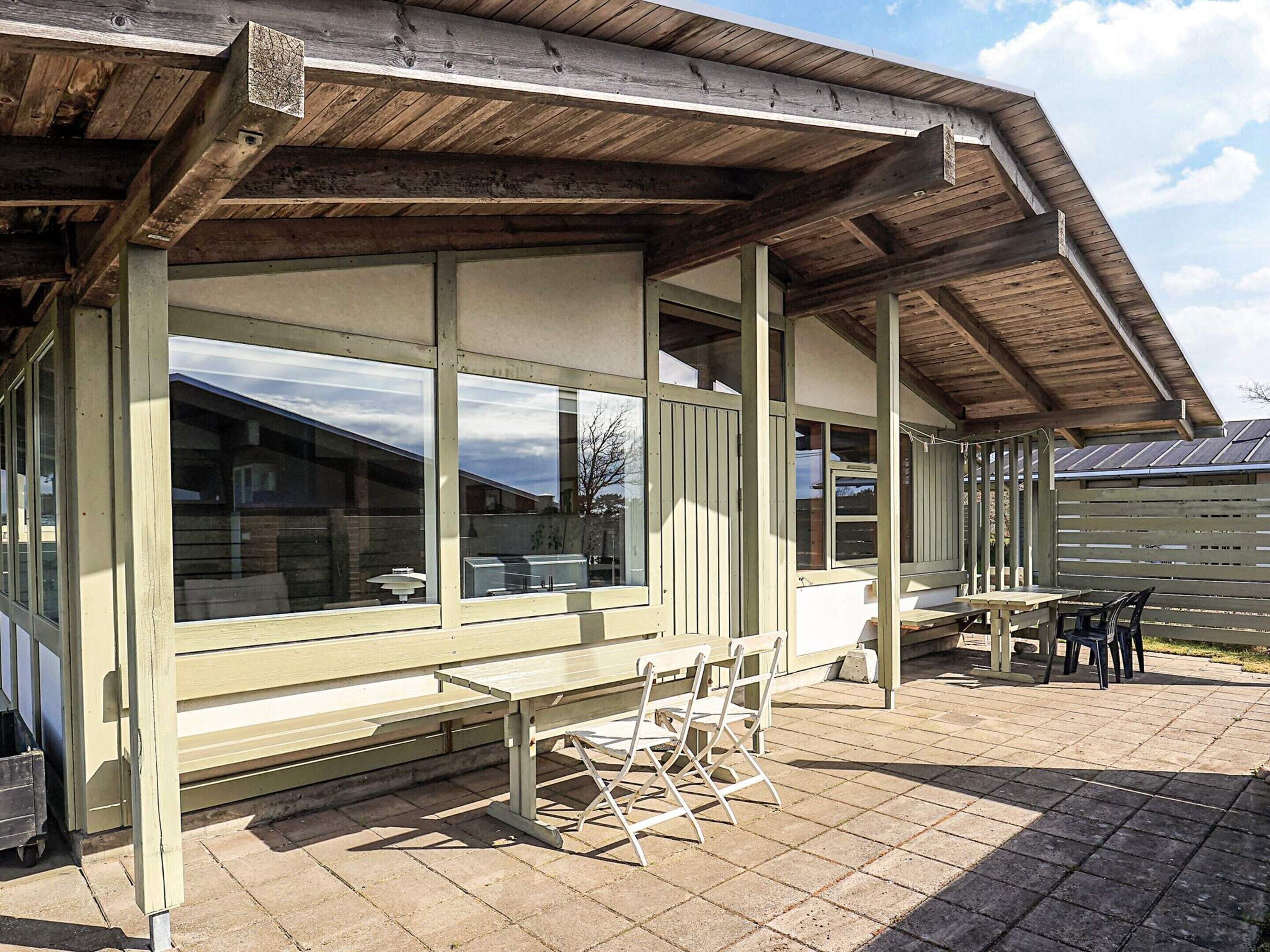 A covered outdoor seating area with a table and chairs, large windows, and a wooden roof structure.