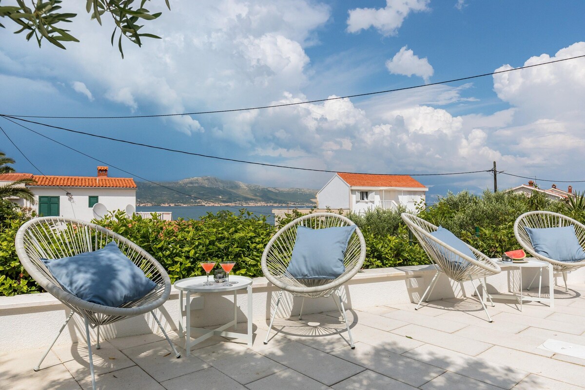 A terrace with wicker chairs and a table, overlooking a scenic view with buildings and greenery.