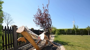 Eine Hängematte an einem gepflasterten Weg, mit einer Holzskulptur, einem Zaun und einer Windkraftanlage im Hintergrund.