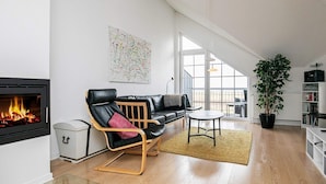 Ett modernt vardagsrum med en öppen spis, en svart lädersoffa, ett träbord och ett stort fönster med utsikt över omgivningen.
