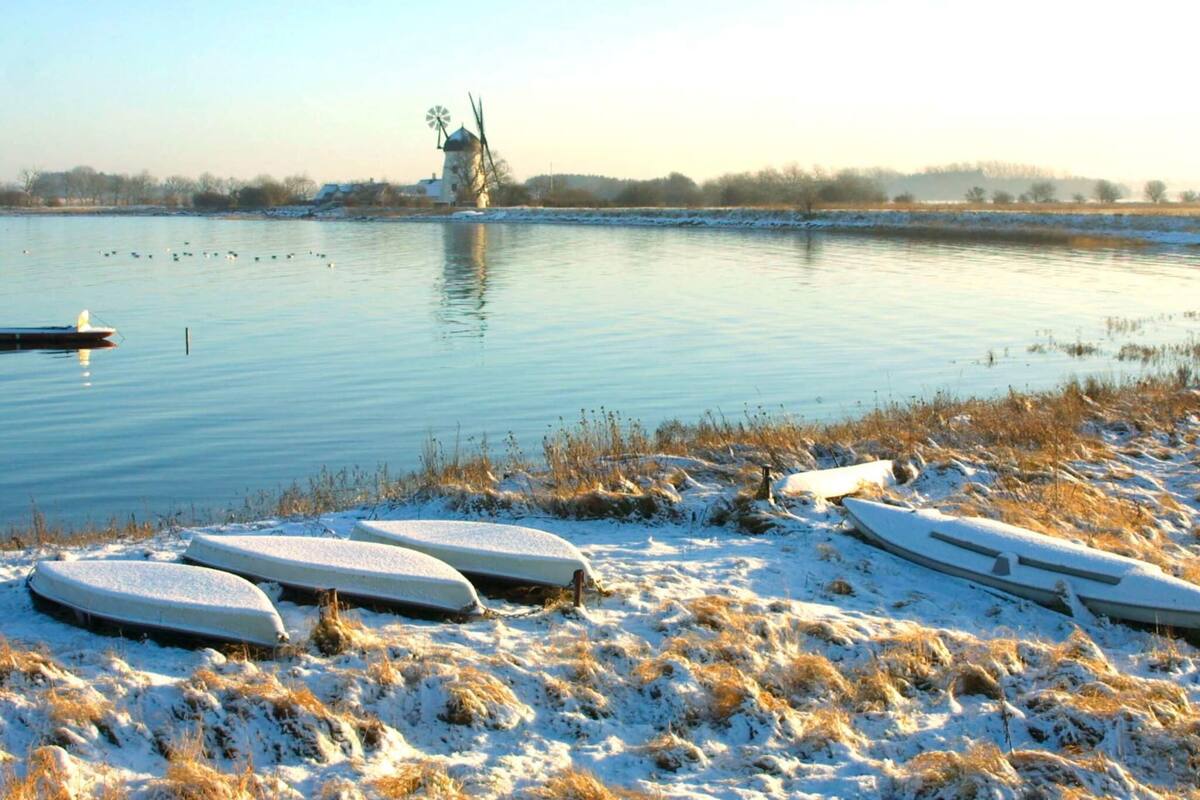 A serene lakeside scene with a windmill in the distance, snow-covered ground, and boats on the shore.