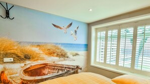 Une chambre avec une fresque représentant une scène de plage, une fenêtre avec des stores et un lit recouvert d’une courtepointe orange.