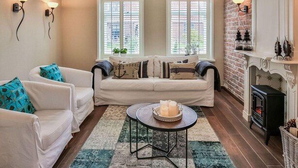 Un salon chaleureux avec une cheminée, deux fenêtres équipées de stores, un canapé blanc agrémenté de coussins à motifs et une table basse décorée de bougies.