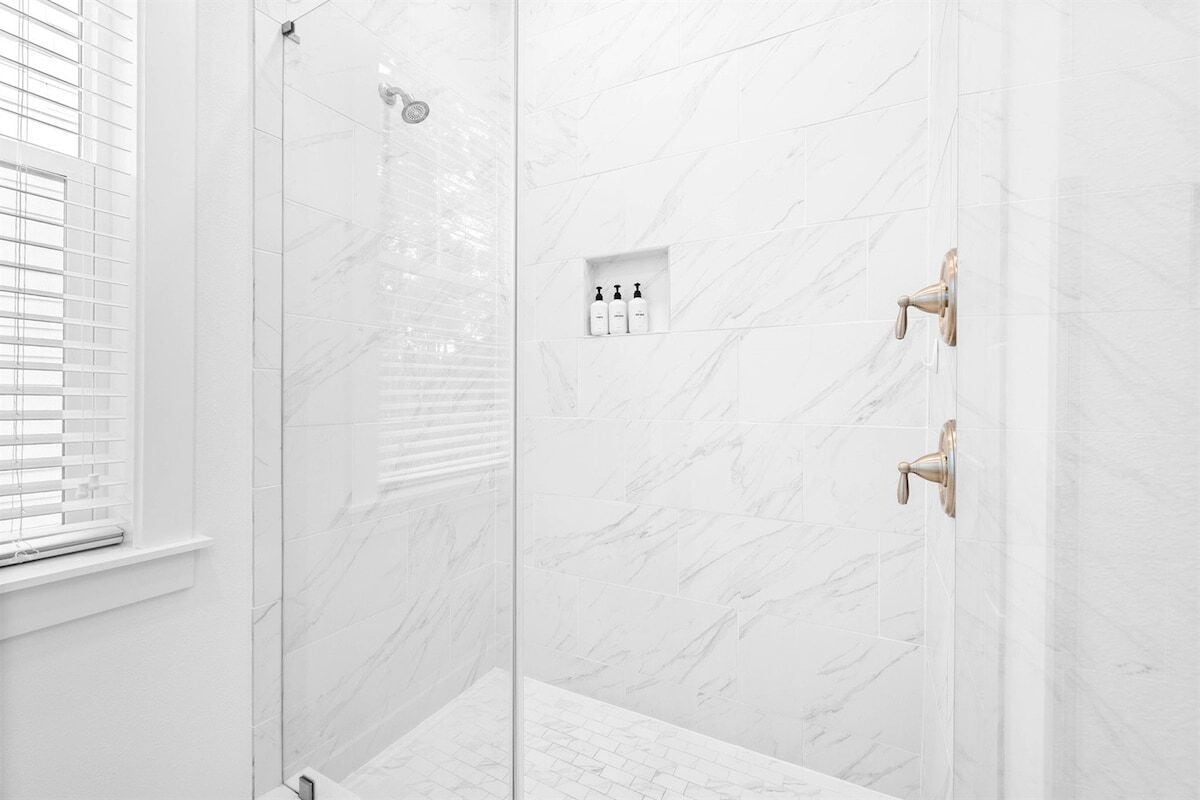A modern bathroom with white marble tiles, a glass shower enclosure, and a window with blinds.