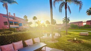 An outdoor seating area with tables and chairs, surrounded by palm trees and a pool, with buildings in the background. - Coastal Apartment/pool/garden/kingbed/acon/freep (Estepona)