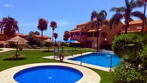 A swimming pool area with a circular pool, palm trees, and a building with a red-tiled roof. - Coastal Apartment/pool/garden/kingbed/acon/freep (Estepona)