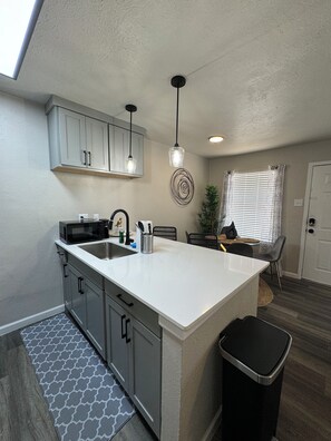A compact kitchen with a white island, grey cabinets, and a black trash bin. - Downtown Dallas Urban Escape (Dallas)
