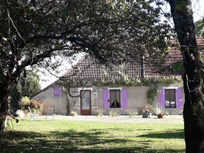 Een landelijk huis met een pannendak, paarse luiken en een tuin met gazon en bloempotten.