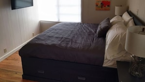 Une chambre à coucher avec un lit, un téléviseur fixé au mur, une fenêtre avec des stores et un tableau accroché au mur.