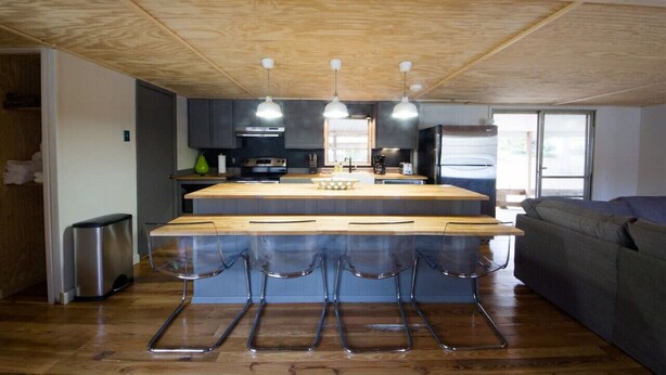 Une cuisine moderne dotée d’un îlot central, de tabourets de bar, de luminaires suspendus et d’un canapé gris dans l’espace de vie adjacent.
