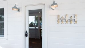 A white house exterior with a front door, two wall-mounted light fixtures, and a decorative metal fence. - Authentic 1940's renovated Folly cottage! (Charleston)