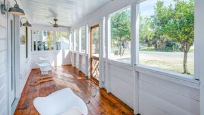 A covered porch with white walls and wooden flooring, featuring a ceiling fan, two white chairs, and a view of a tree-lined street. - Authentic 1940's renovated Folly cottage! (Charleston)