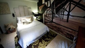 A bedroom with a bed, a nightstand, a lamp, a wall-mounted shelf, and a large abstract painting. - Studio- Behind Bert’s Market (Folly Beach)