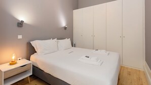 Egy gondosan megterített ágy fehér ágyneművel, egy éjjeliszekrény lámpával, és egy tolóajtós szekrény.