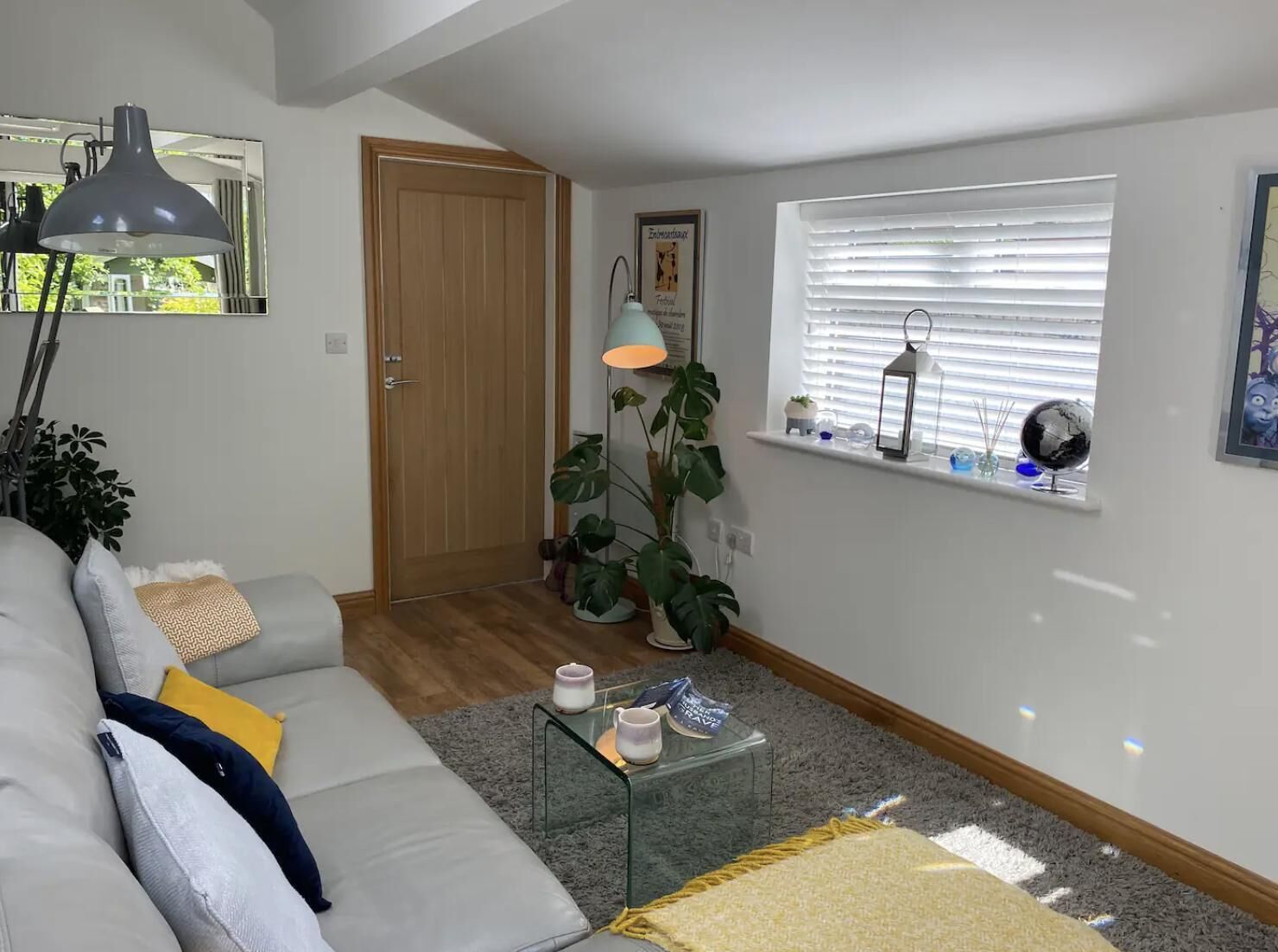 Un salon comprenant un canapé, une table basse en verre, une lampe, une porte et une fenêtre avec des stores.