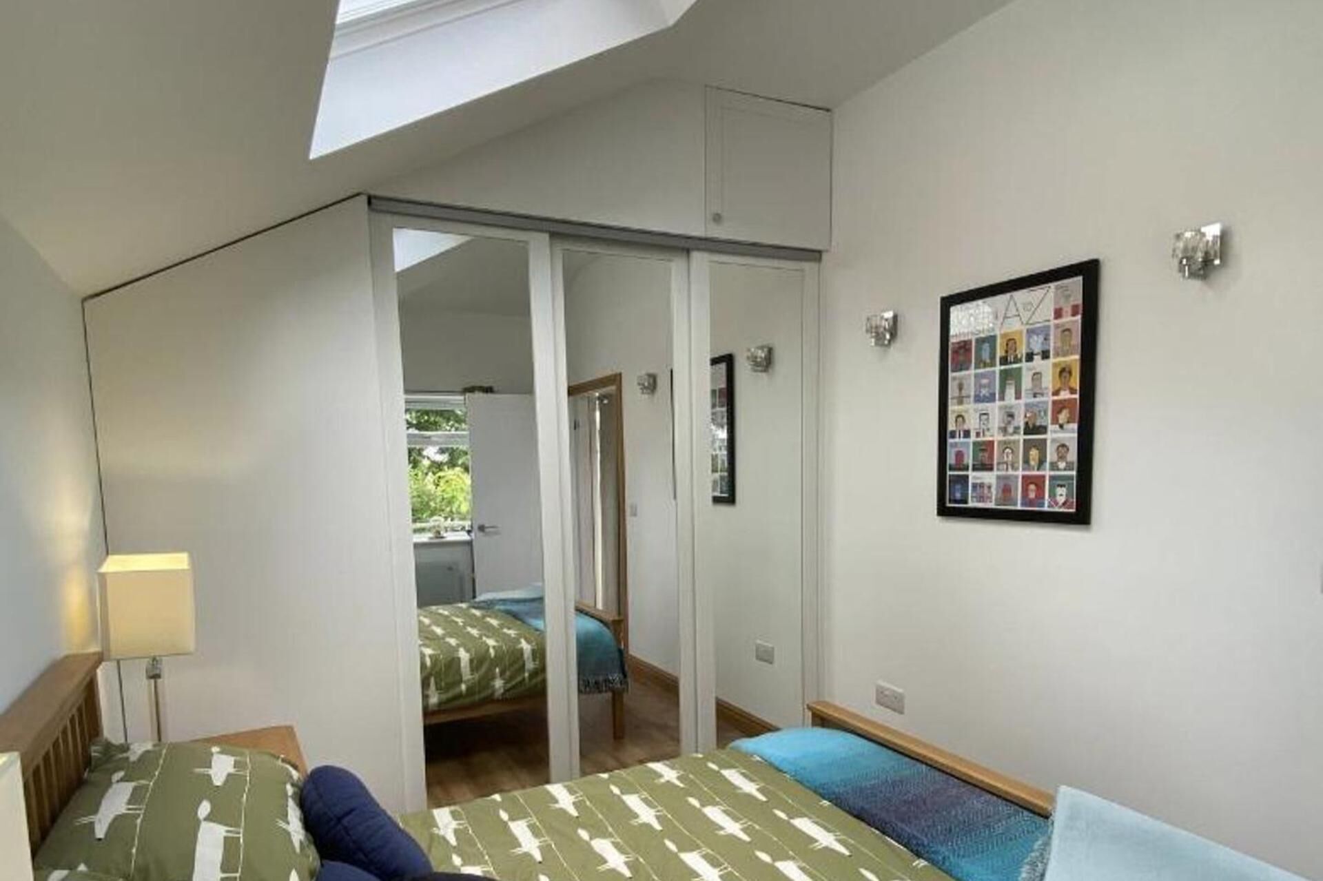 Une chambre dotée d’une lucarne, d’un lit avec une literie à motifs, d’une table de chevet, d’une commode, d’un mur orné d’un collage de photos encadrées et d’une porte coulissante donnant sur une autre pièce.