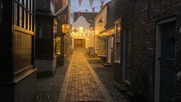 Une ruelle étroite pavée de pierres, bordée de bâtiments des deux côtés, avec un mur de briques sur la droite et, au loin, une enseigne indiquant le pub « The George Morton ».