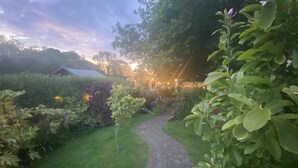 Un sentier de jardin mène à un bâtiment doté d’une zone abritée, le tout étant entouré d’une végétation luxuriante et de plantes en fleurs.