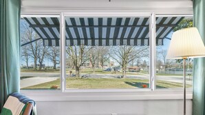 A room with a view of a park, a green cushioned bench, a lamp, and a window with a striped awning. - Walk to Lambeau! Sleeps 8• Fire Pit • Party Garage • Parking for 6 vehicles (Ashwaubenon)