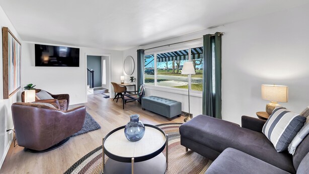 A modern living room with a sofa, armchair, and round coffee table. There is a TV mounted on the wall, a lamp, and a view of the outdoors through large windows. - Walk to Lambeau! Sleeps 8• Fire Pit • Party Garage • Parking for 6 vehicles (Ashwaubenon)