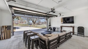 A spacious garage with a large opening, a sofa, bar stools, a flat-screen TV, and a projector mounted on the ceiling. - Walk to Lambeau! Sleeps 8• Fire Pit • Party Garage • Parking for 6 vehicles (Ashwaubenon)