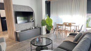 A modern living room with a grey sofa, a coffee table, and a TV mounted on the wall. There is a large plant, a dining area with wooden chairs, and a large window with curtains.