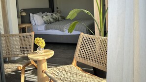 A room with a bed, two wicker chairs, a small round table with a vase of flowers, and a large plant.