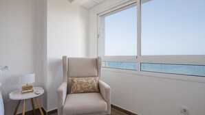 Een kamer met uitzicht op de zee, een stoel met kussen, een klein tafeltje met lamp, en een stopcontact aan de muur.