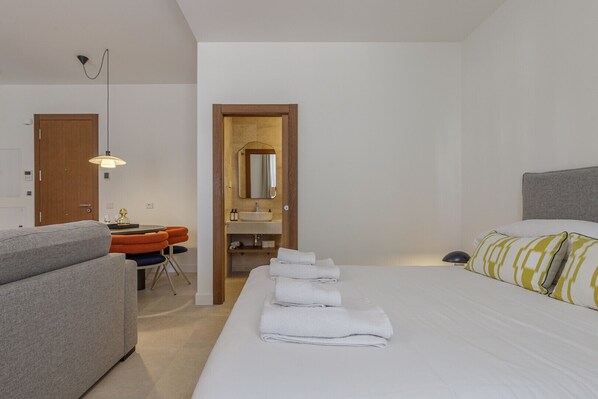 Habitación de hotel con cama, escritorio con silla, baño con lavamanos y espejo, y una puerta.