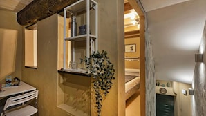 Une pièce de taille modeste comprenant un lit, une petite table avec des chaises, une étagère avec quelques objets et une fenêtre agrémentée d’une plante.