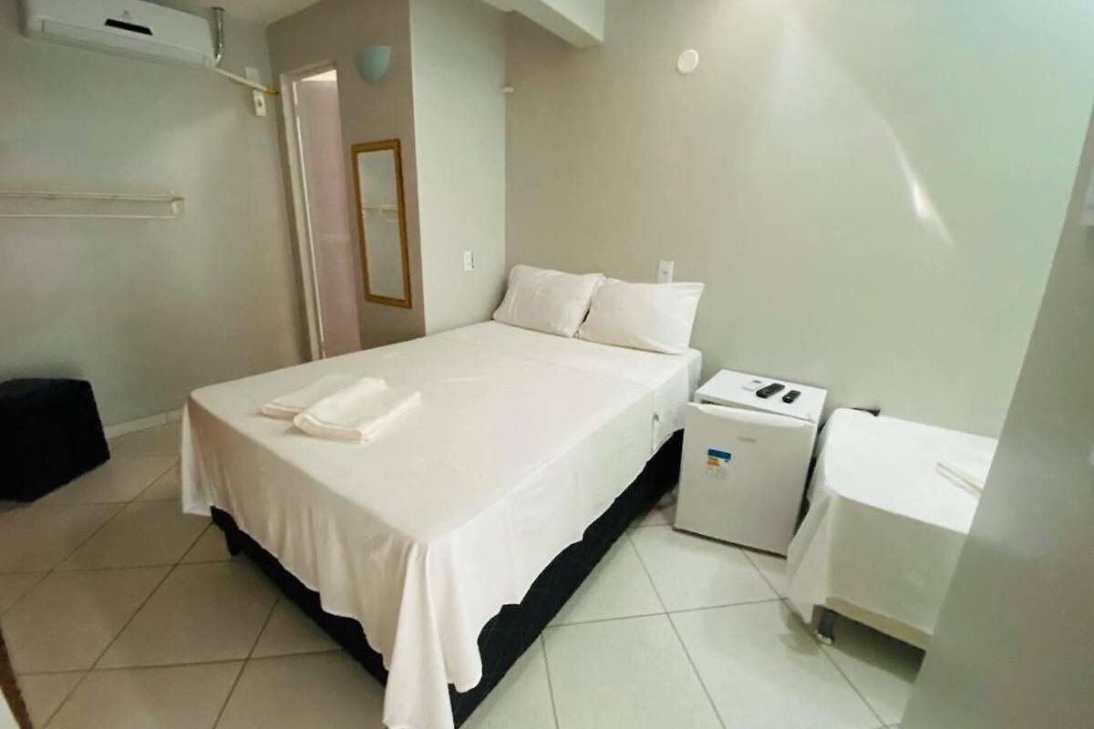 A clean, modern hotel room with a neatly made bed, a bedside table, a small desk, and a chair.