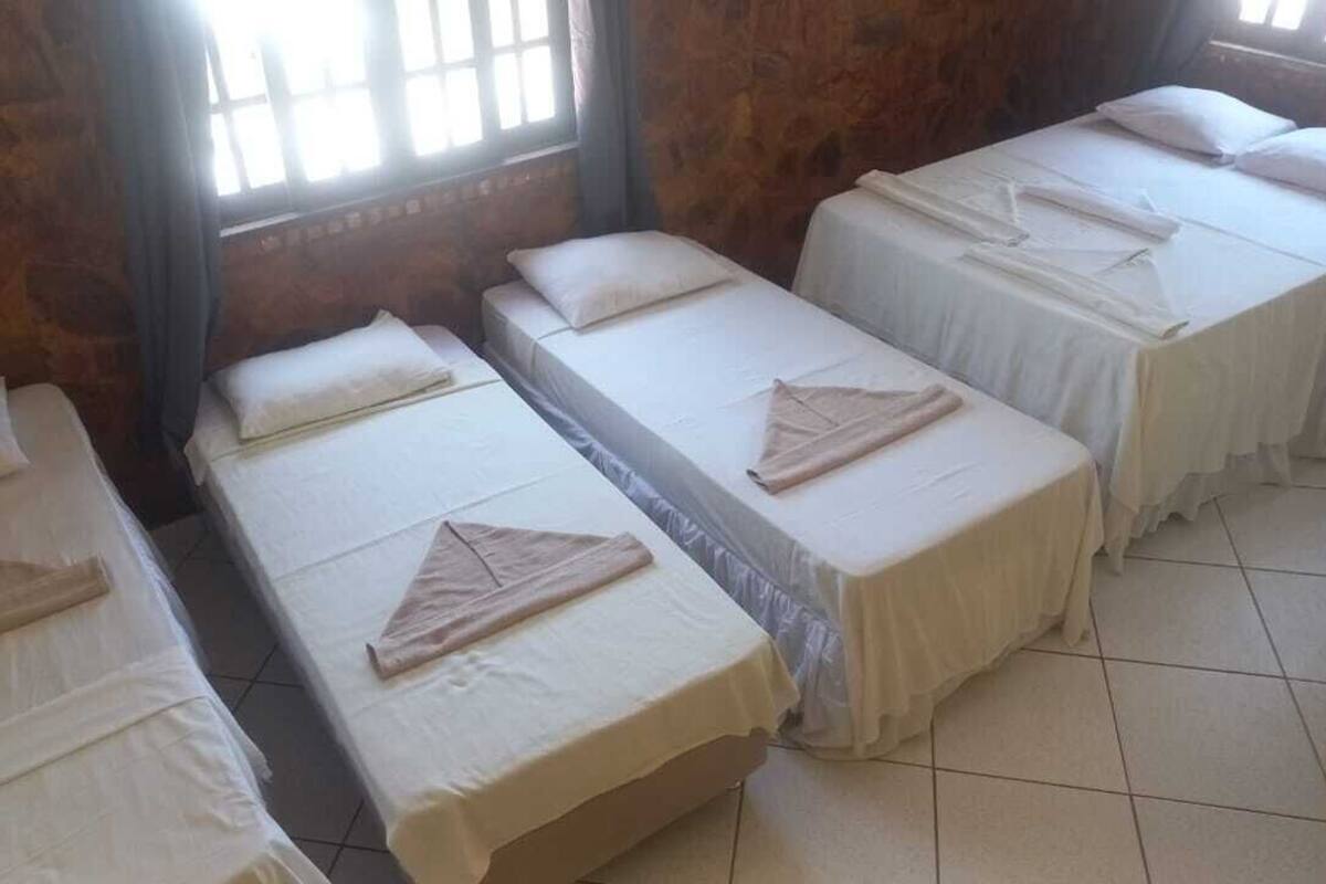 A room with four single beds arranged in a row, each with a white bedspread and a folded towel. The room has stone walls and a window with curtains.
