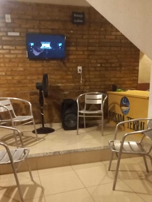 A room with a brick wall, a television mounted above a desk, and several metal chairs. - Pousada Terral (Aracati)