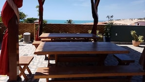 Une terrasse extérieure avec des tables et des bancs en bois, un mur de briques et une vue sur la plage et l’océan.