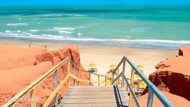 Un escalier en bois mène à une plage de sable fin bordée d’une eau turquoise et cristalline.