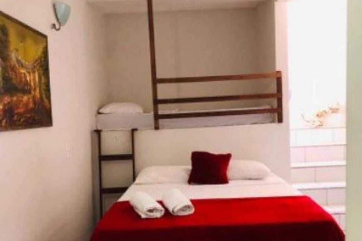 A small, single-room hotel room with a bunk bed, a red bedspread, white curtains, and a painting on the wall.