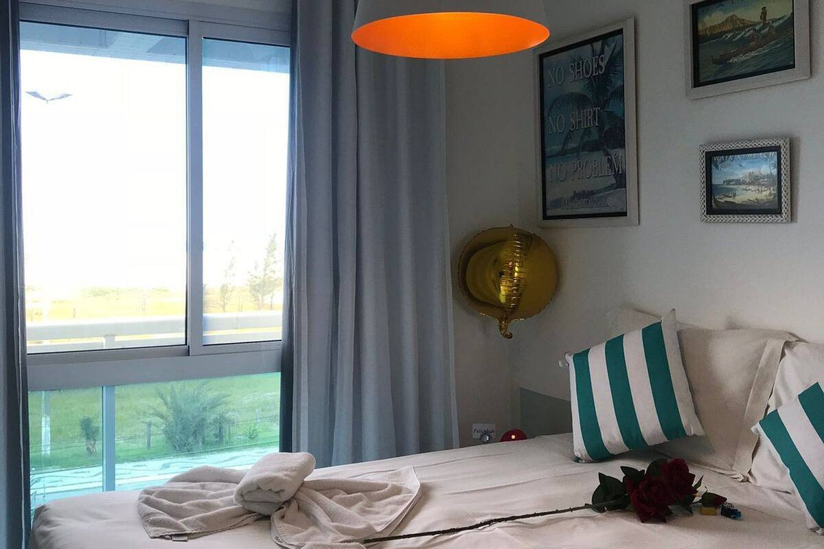 A hotel room with a bed, a large window, a hanging lamp, and framed pictures on the wall.