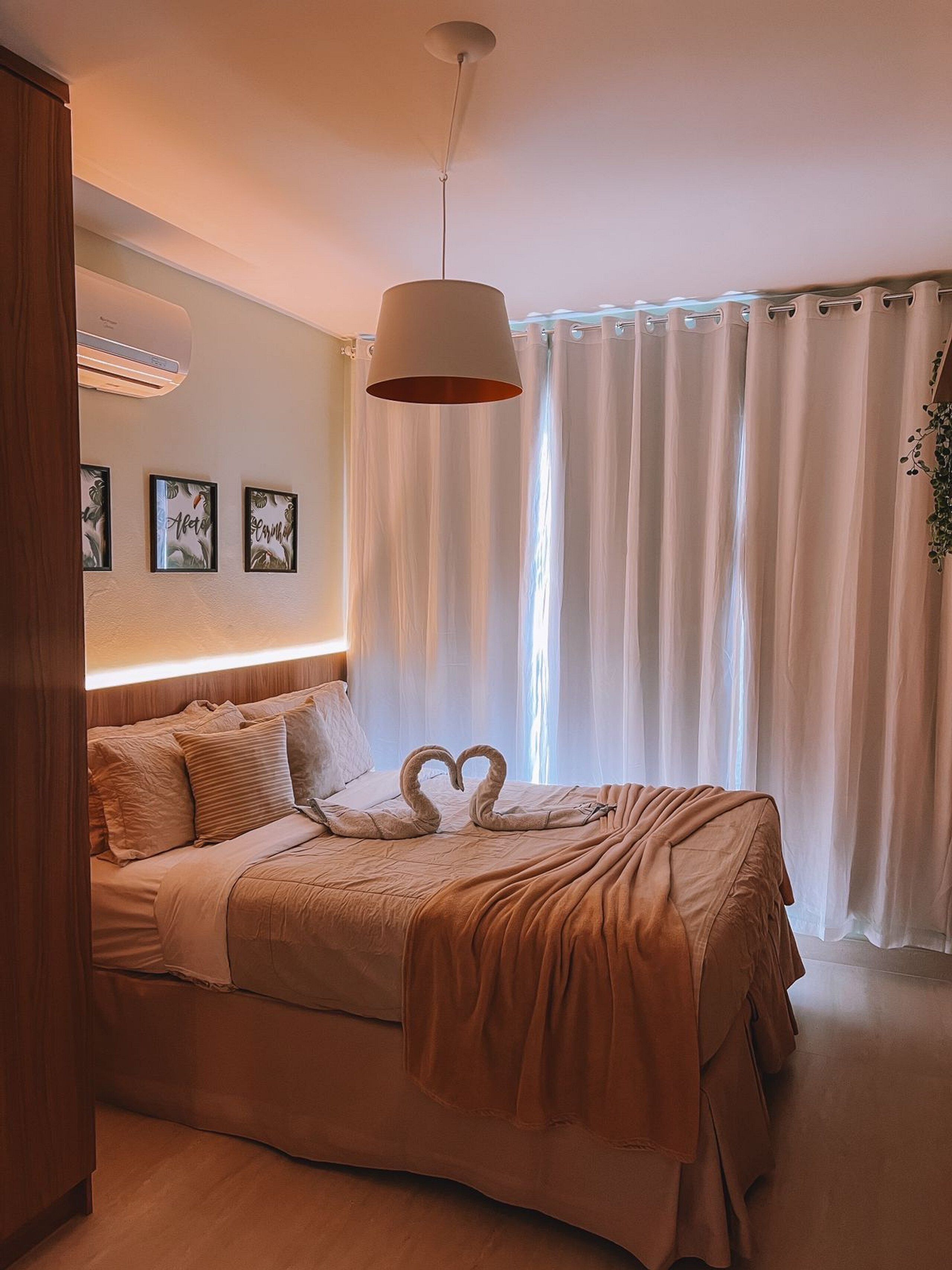 Une chambre à coucher comprenant un lit, un luminaire suspendu, un climatiseur fixé au mur et une fenêtre avec des rideaux.