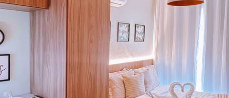A hotel room with a wooden wardrobe, a bed with a heart-shaped towel arrangement, a bedside table with a microwave, and a hanging lamp.